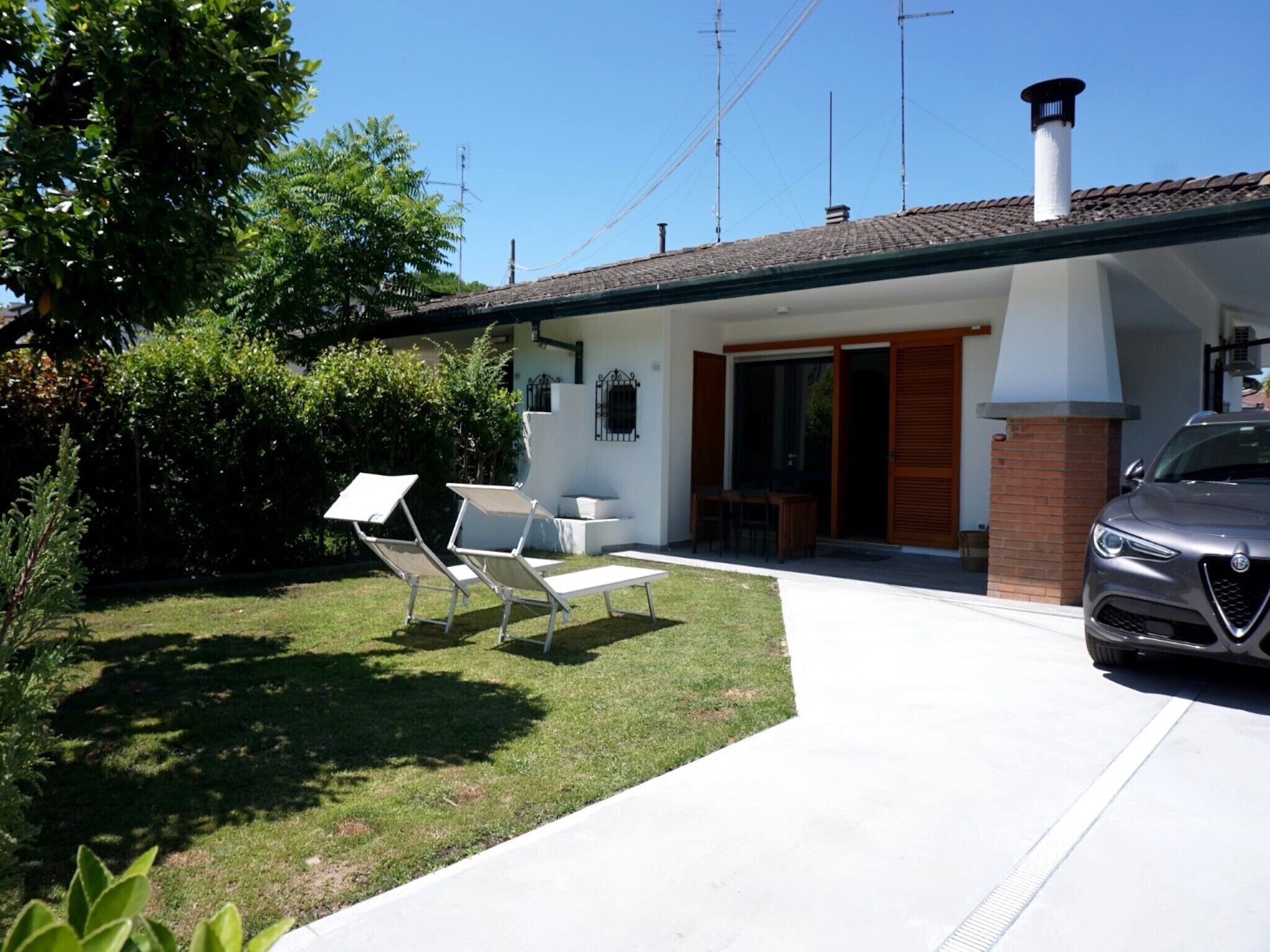 Una casa moderna con garaje, un jardín con dos sillones blancos y un coche aparcado.