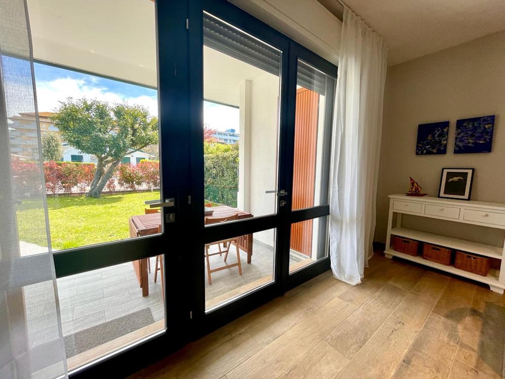 A room with a sliding glass door leading to a patio with a table and chairs, a white console table with drawers, and a view of a garden and buildings outside.