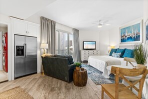 Una habitación de hotel moderna con una cama grande, un sofá, una silla de madera, un televisor y una zona de cocina con refrigerador y lavadora.