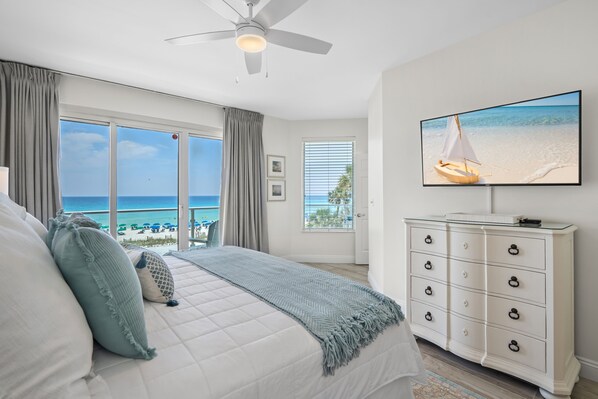 Un dormitorio con una cama grande, ventilador de techo, televisor en la pared y vista a la playa.