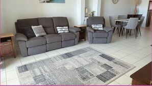Una sala de estar con un sofá gris, sillones, una mesa de comedor con sillas, un tapiz y un cuadro en la pared.