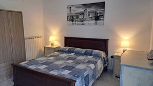 Une chambre à coucher avec un lit en bois, deux tables de chevet avec des lampes, un radiateur, une armoire et un tableau représentant un paysage urbain accroché au mur.