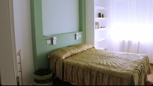 Un dormitorio con cama, una mesita de noche, un estante en la pared y una ventana con cortinas.