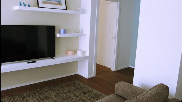 Una sala de estar con un televisor de pantalla plana, estanterías blancas, un sofá marrón y una puerta que da a otra habitación.