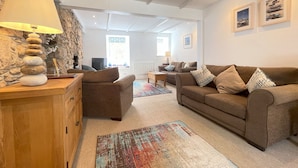 A cozy living room with a stone wall, a wooden cabinet, a brown sofa, and a colorful rug. - Howards Way - a cottage that sleeps 5 guests in 2 bedrooms (St Ives)