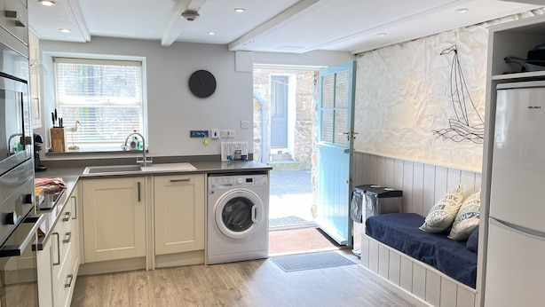 A compact kitchen with a washing machine, a sink, and a window with blinds. - Howards Way - a cottage that sleeps 5 guests in 2 bedrooms (St Ives)