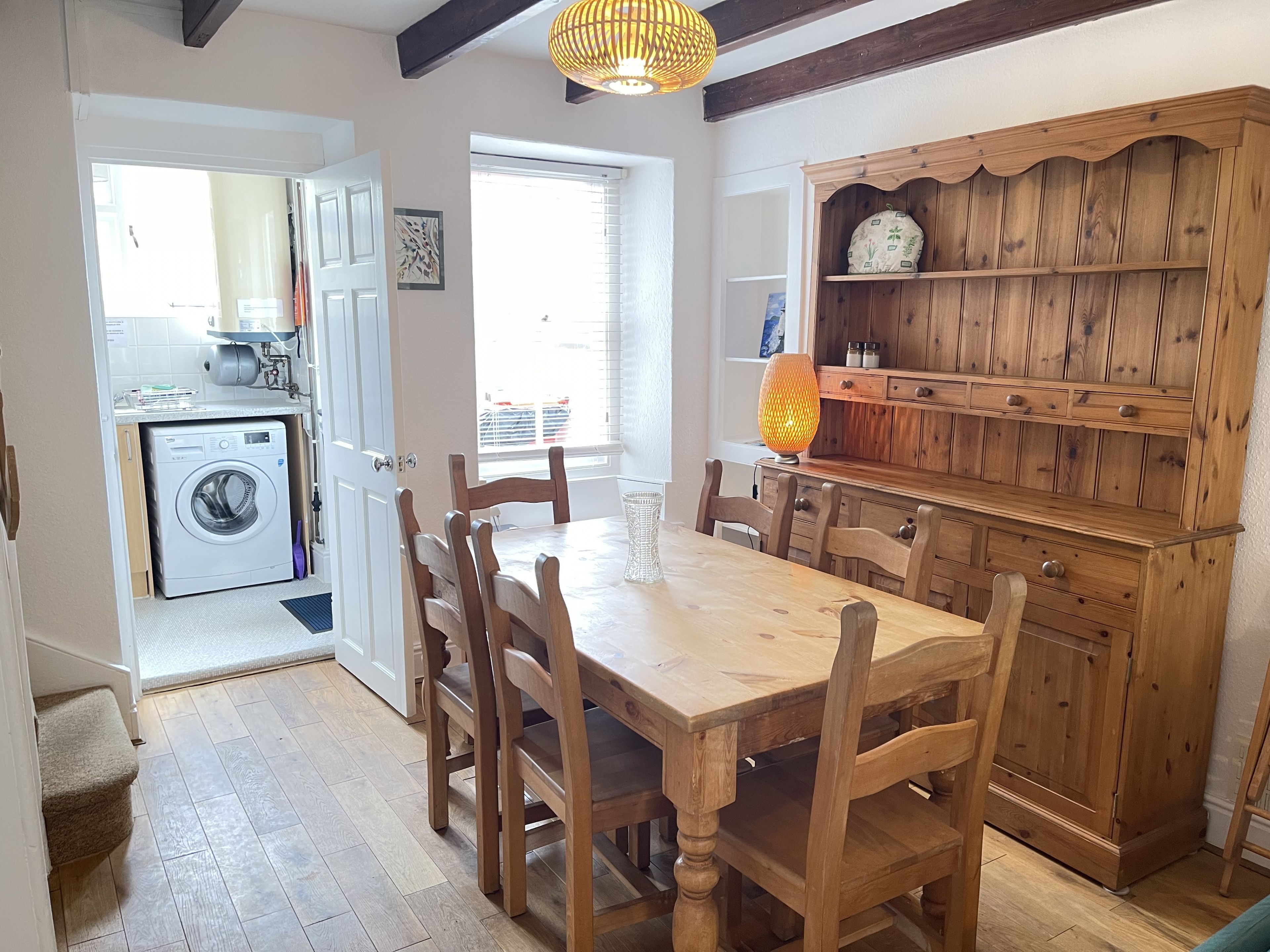 Een eetruimte met een houten tafel en stoelen, een houten kast met planken, en een wasmachine in de aangrenzende kamer.