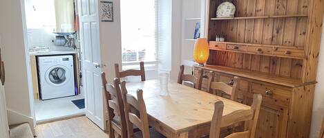 Een eetruimte met een houten tafel en stoelen, een houten kast met planken, en een wasmachine in de aangrenzende kamer.