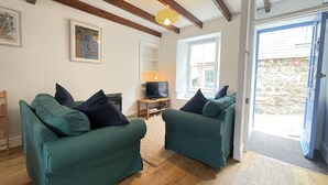 A living room with two green sofas, a wooden coffee table, a TV, and a window with a view of a stone wall. - Silver Seahorse - a cottage that sleeps 6 guests in 3 bedrooms (St Ives)