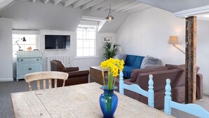 Un espace de vie de style loft avec un plafond haut, des poutres apparentes, une table à manger en bois, un vase bleu avec des fleurs, un canapé en cuir marron et un divan à motifs bleus et blancs.