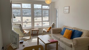 Ein Wohnzimmer mit einem beigefarbenen Sofa, einem Holztisch und einem Computertisch. Große Fenster bieten Blick auf einen Strand und Boote.