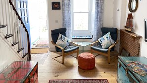 A cozy living room with a staircase, two armchairs, a small ottoman, a glass-top table, and a blue trunk. - Sea Pebble - a cottage that sleeps 4 guests in 2 bedrooms (St Ives)