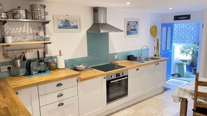 A modern kitchen with a built-in oven, a sink, and a stove. There are shelves with kitchenware and a window with a view of the outside. - Sea Pebble - a cottage that sleeps 4 guests in 2 bedrooms (St Ives)