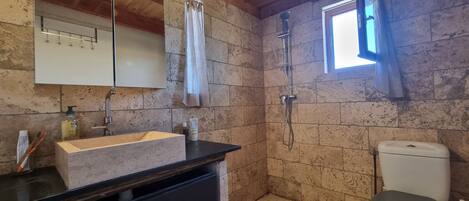 A bathroom with a stone wall, a sink with a mirror, a shower, and a toilet.