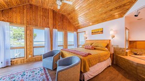 Quarto com teto de madeira, grandes janelas com vista para o mar, cama com colcha estampada, duas poltronas e uma mesinha.