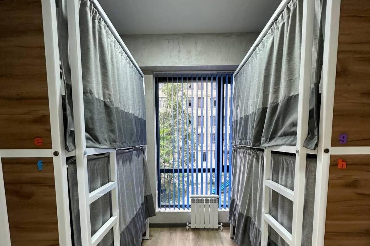 A narrow corridor with bunk beds on either side, wooden flooring, and a view of a building through a barred window.