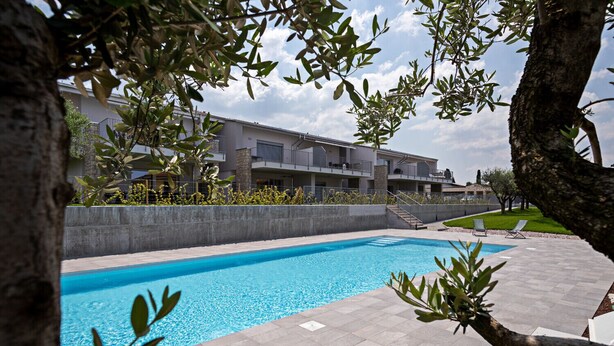 Une piscine dotée d’une plage en béton, entourée d’un petit muret en pierre, et un bâtiment avec des balcons en arrière-plan.