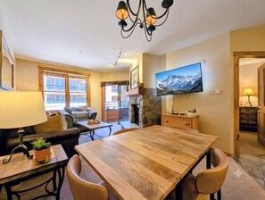 A cozy living room with a wooden dining table, comfortable seating, a TV mounted on the wall, and a fireplace. - Courtyard Views, Walk to Gondola, On-site Hot Tub (Keystone)