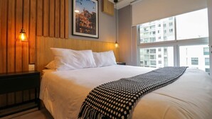 Une chambre d’hôtel avec un grand lit, un mur lambrissé en bois, un tableau encadré et une fenêtre donnant sur des immeubles.