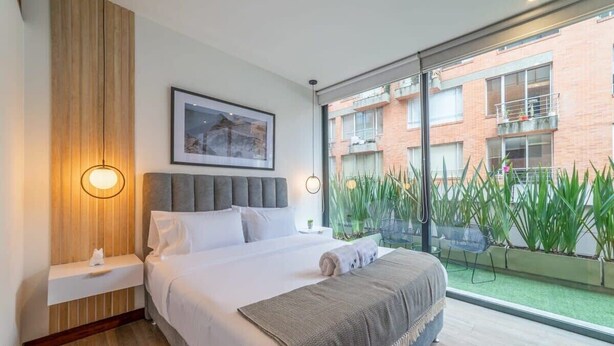 Ein modernes Hotelzimmer mit einem großen Bett, einer Wand mit Holzverkleidung, einem gerahmten Bild und Blick auf ein Backsteingebäude sowie Grünpflanzen durch große Fenster.