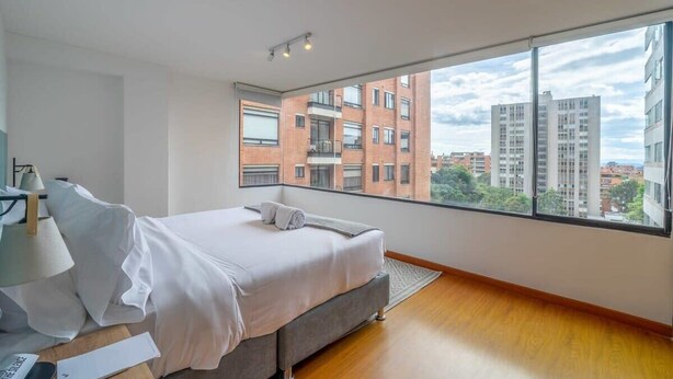 Un dormitorio moderno con un ventanal grande, una cama, una mesita de noche con una lámpara y vistas de edificios circundantes.