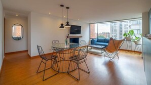Una sala moderna con una mesa redonda para comer, sillas negras de estructura de alambre, un sofá azul y un televisor montado en la pared.