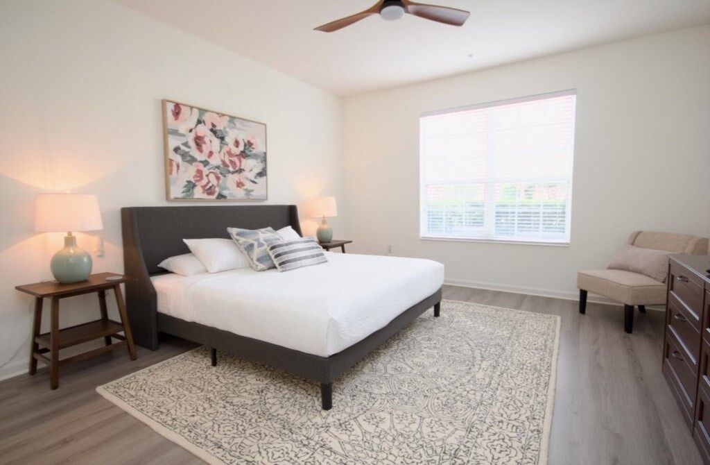 A bedroom with a bed, bedside lamps, a ceiling fan, a window, and a dresser.