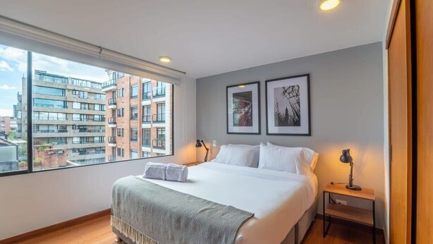 A modern hotel room with a large bed, two bedside tables, a window with a city view, and framed artwork on the wall. - w* | Industrial 1BR In Cabrera (Bogotá)