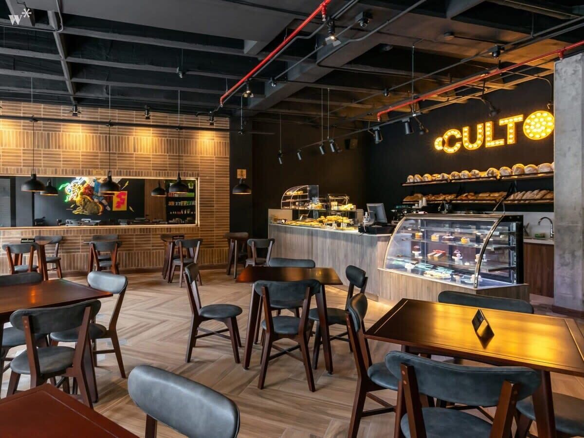 Een modern caféinterieur met houten vloeren, zwart-grijze stoelen, een balie met een vitrine en een neonbord met de tekst 'CULTO'.