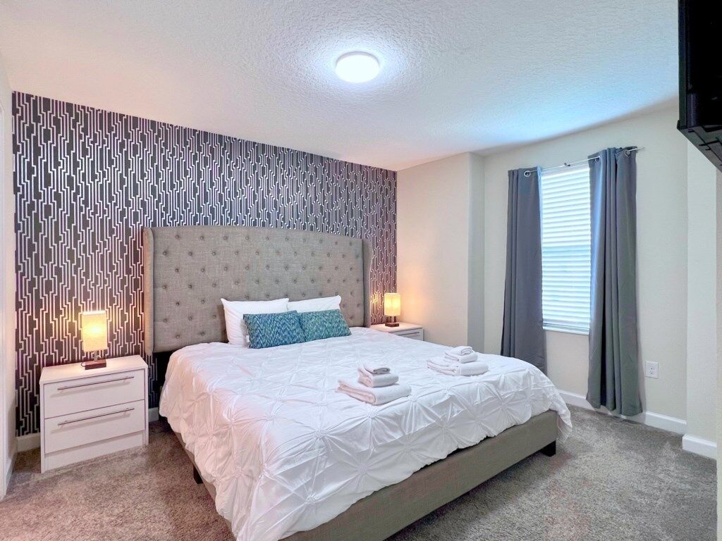 Een keurig opgemaakte slaapkamer met een groot bed, twee nachtkastjes, een behang met patroon en een raam met gordijnen.