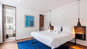 Ein modernes Hotelzimmer mit einem großen Bett, zwei Nachttischen mit Lampen, einem blauen Teppich, einem gerahmten Kunstwerk an der Wand und Blick ins Badezimmer durch eine geöffnete Tür.