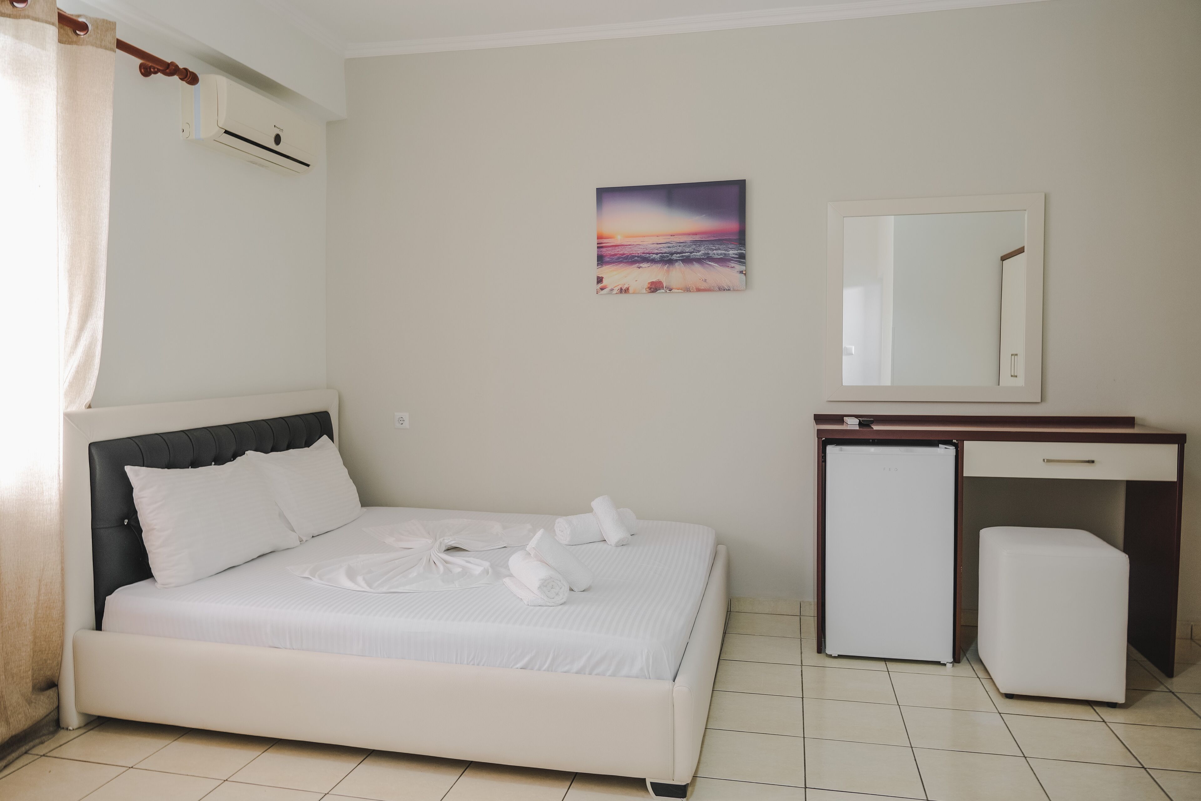 A bedroom with a bed, a small refrigerator, a mirror, and a painting of a sunset on the wall.