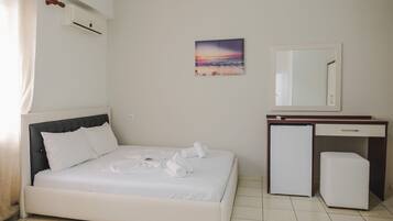 A bedroom with a bed, a small refrigerator, a mirror, and a painting of a sunset on the wall.