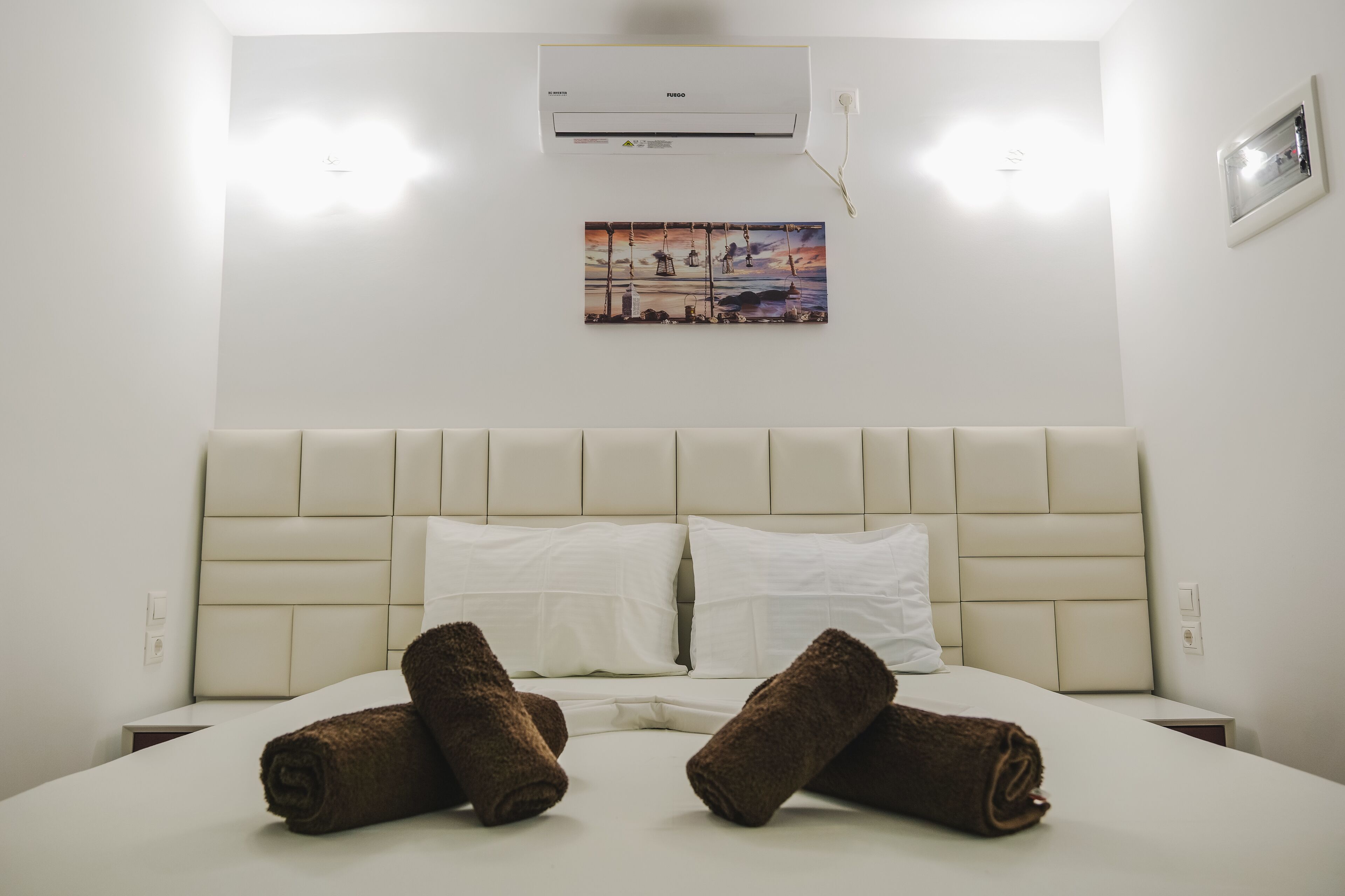 Una habitación de hotel con una cama grande, cabecera blanca, dos almohadas blancas y dos toallas enrolladas.