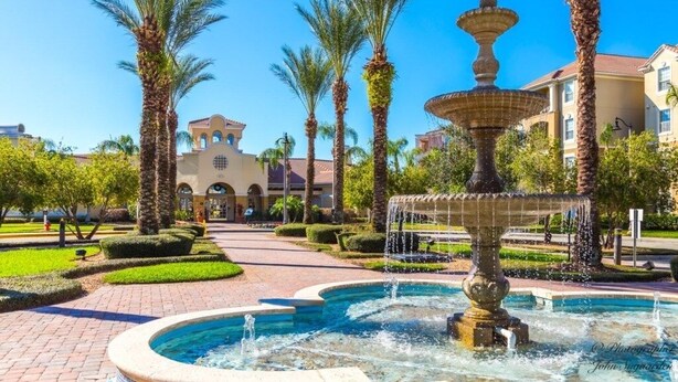 A fountain with water jets in a landscaped area with palm trees and a building with arched windows in the background.