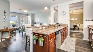 A modern kitchen with granite countertops, wooden cabinets, and a central island. There is a dining area with a table and chairs, and a living room with a sofa and coffee table.