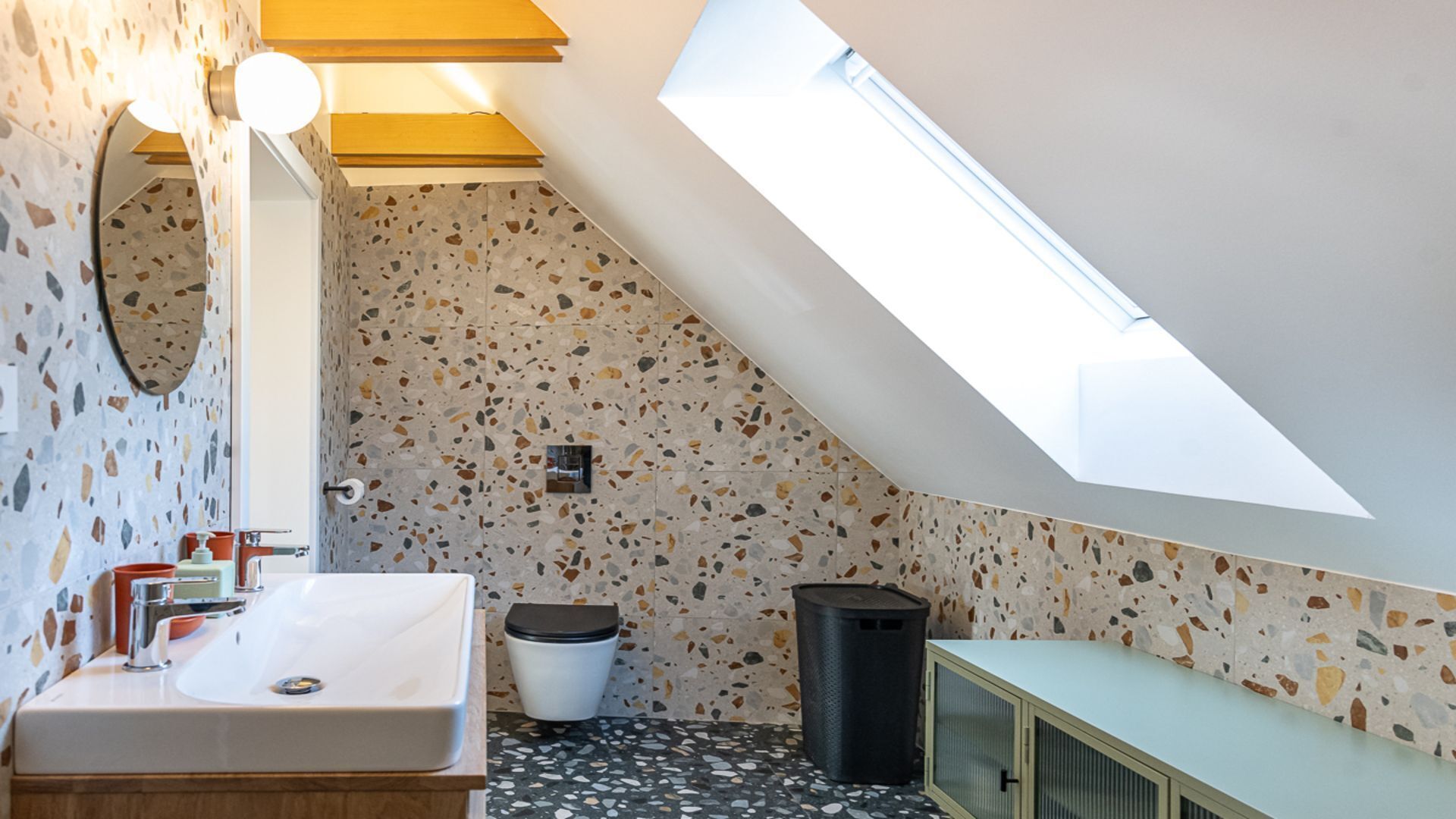 Un bagno moderno con finestra sul tetto, pavimento in terrazzo, lavabo bianco e mobile.