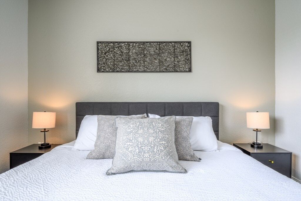 A neatly made bed with a grey headboard, two bedside tables with lamps, and a wall-mounted abstract art piece.