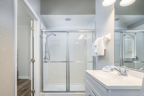 Un baño moderno con ducha de mampara de vidrio, tocador blanco con lavamanos y un espejo grande.
