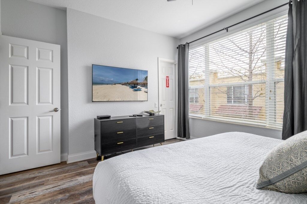 Ein Schlafzimmer mit einem großen Bett, einer Kommode, einem Bild von einem Strand und einem Fenster mit Jalousien.