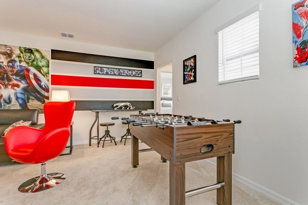 A room with a foosball table, a red chair, a sofa, and a wall with a superhero mural.
