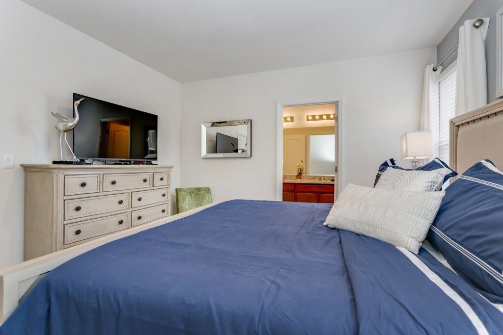 A bedroom with a large bed, a dresser, a television, a mirror, and a vanity with lights.