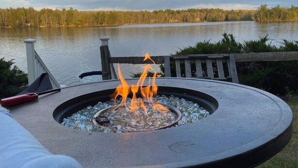 A circular outdoor fire pit with flames, surrounded by a metal railing, overlooking a lake with trees in the background.