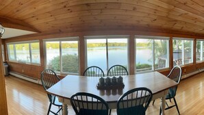 A dining area with a wooden table and chairs, large windows with a view of a lake and greenery.