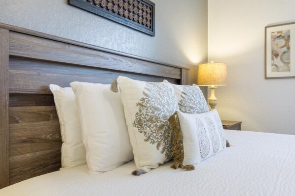 A neatly made bed with white bedding and decorative pillows, a wooden headboard, a bedside lamp, and a framed artwork on the wall.