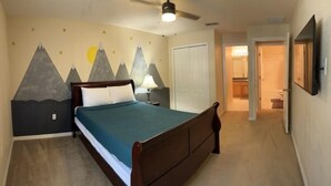 Un dormitorio con cama, ventilador de techo, pared decorada con motivos de montaña, una puerta y televisor montado en la pared.