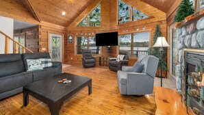 Amplia sala de estar con techo alto, paredes de madera y ventanas grandes. Hay una chimenea, un televisor y un área de descanso con un sofá y sillones.