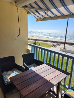Balcón con mobiliario de mimbre y una mesa de madera, con vistas a una playa y al océano.
