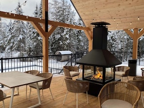 A covered outdoor seating area with a wood-burning stove, wicker chairs, and a view of snow-covered trees.