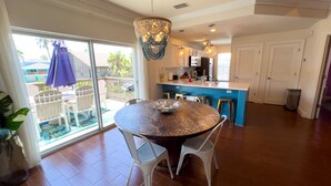 A spacious dining area with a round table and chairs, a kitchen in the background, and a view of a pool through a sliding glass door. - Sun Dancer #11 2 Bedroom Home by RedAwning (South Padre Island)